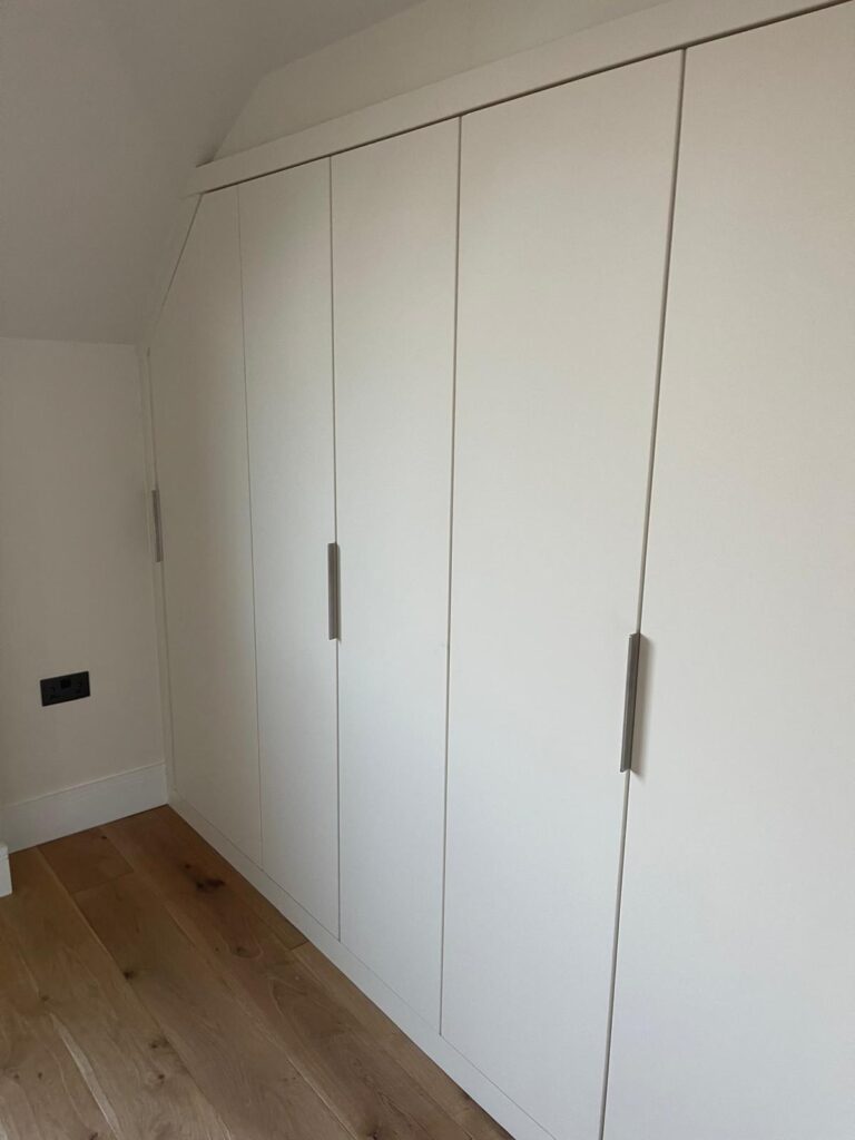 Floor-to-ceiling white fitted wardrobes with brushed steel handles, built to a sloping ceiling on an oak floor, by R3 Carpentry