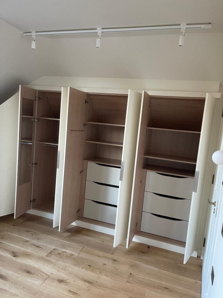 White fitted wardrobe under a sloping loft ceiling with oak interior, drawers, shelves and hanging rails, by R3 Carpentry